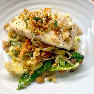 A piece of white fish topped with crispy breadcrumbs sits on a bed of mixed vegetables and pasta, served on a white plate.