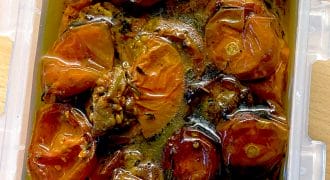 A plastic container filled with slow-roasted tomatoes covered in olive oil, showing shrivelled, glossy red tomato halves and seasoning.