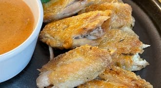 Crispy baked chicken wings arranged on a black plate, served with a pot of orange dipping sauce and a garnish of fresh coriander.