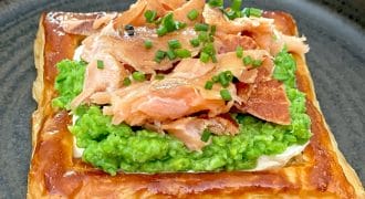 A square puff pastry topped with a layer of mashed peas, flaked smoked salmon, and chopped chives, served on a dark round plate.