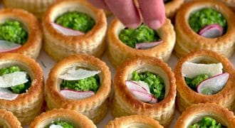 A hand garnishes round puff pastry shells filled with garden peas, topped with radish slices and cheese shavings, arranged neatly on a tray.