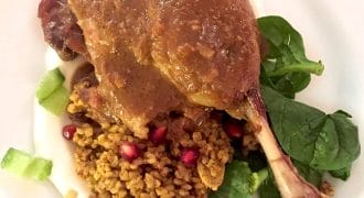 A cooked duck leg with brown sauce is served on a bed of spinach, bulgur or couscous, pomegranate seeds, and diced celery, plated on a white plate.