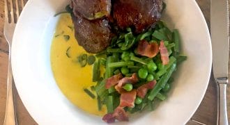 A white plate with three pieces of glazed meat, green beans and peas topped with bacon pieces, and yellow sauce, set on a wooden table with a fork and knife beside the plate.