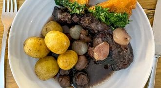 A plate with boiled baby potatoes, pearl onions, mushrooms, braised meat in dark sauce, and two triangular pieces of toast topped with chopped herbs. Knife and fork are placed on either side of the plate.