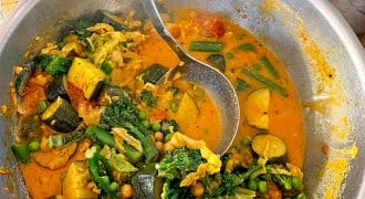 A large metal bowl filled with vibrant vegetable curry, including courgette, broccoli, peas, green beans, and chickpeas in a rich orange sauce, with a ladle resting inside. A bowl of white rice is in the background.