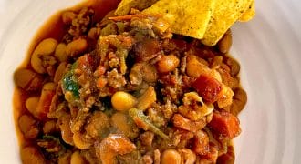 A plate of chilli with beans, minced meat, tomatoes, and vegetables, topped with three yellow corn tortilla crisps on the side. The chilli appears hearty and saucy, served on a white plate.