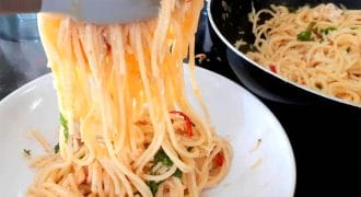 A close-up of spaghetti being served from a frying pan into a white bowl using kitchen tongs. The pasta is mixed with herbs and sliced red chilli peppers.
