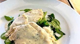 Two pieces of chicken breast in a creamy white herb sauce served on a bed of fresh spinach leaves on a white plate, with a bowl of whole baby potatoes in the background.