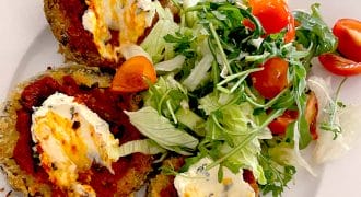 Three breaded aubergine slices topped with tomato sauce and melted cheese are served on a white plate with a side salad of mixed leaves and cherry tomatoes.