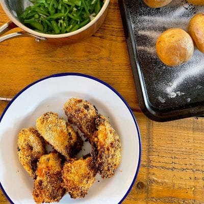 A plate of crispy breaded chicken pieces, a saucepan of green beans, and a baking tray with baked potatoes on a wooden table.
