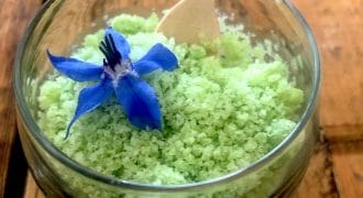 A glass beaker filled with light green granulated pudding, topped with a vibrant blue edible flower, and a small wooden spoon resting inside. The beaker sits on a rustic wooden table.