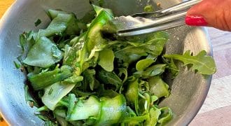 A hand with red nail varnish holds tongs, tossing a green salad with cucumber ribbons and leafy greens in a metal bowl on a wooden surface.