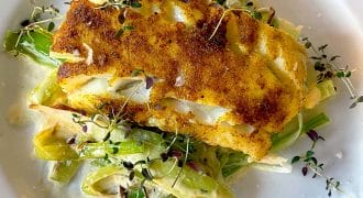 A golden-brown, pan-seared fish fillet sits atop a creamy medley of sautéed vegetables, garnished with fresh microgreens, on a white plate.