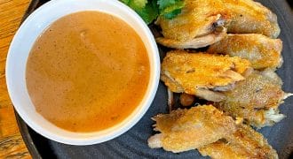 A black plate with crispy chicken wings, a garnish of coriander, and a white bowl filled with brown dipping sauce, set on a wooden table.