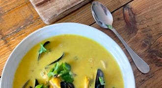 A white bowl of yellow seafood soup garnished with coriander sits on a wooden table next to a metal spoon and a chopping board with a piece of crusty bread. Black mussel shells are visible in the soup.