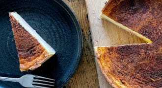 A slice of custard tart on a black plate with a fork beside it sits next to the rest of the tart, which is on a wooden chopping board with one slice missing. The surface of the tart appears caramelised.