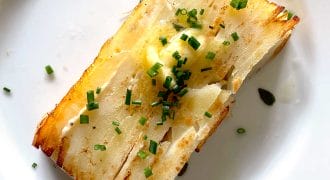 A rectangular slice of layered, golden-brown potato garnished with chopped chives and a pat of melted butter, served on a white plate.