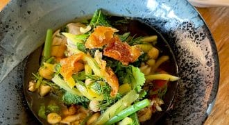 A bowl of vegetable soup garnished with fresh herbs and crispy toppings sits on a wooden table, with a decorative spoon and glass visible nearby.