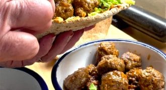 A hand holds a pitta filled with meatballs, lettuce, and sauce. Below, a bowl contains more saucy meatballs, and a small bowl of chopped herbs is nearby on a wooden worktop.
