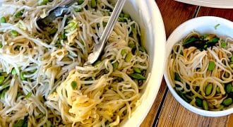 A large white bowl of noodle salad with green vegetables and metal tongs sits on a wooden table next to a smaller bowl of salad and a plate of sliced beef over greens.