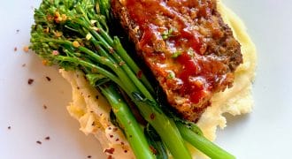 A slice of glazed meatloaf sits on creamy mashed potatoes, garnished with steamed tenderstem broccoli and sprinkled with red chilli flakes, served on a white plate.