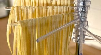 Freshly made pasta noodles hanging to dry on a clear plastic drying rack in a kitchen setting. The uncooked pasta strands are neatly arranged in rows.