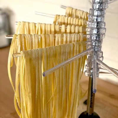 Freshly made pasta noodles hanging to dry on a clear plastic drying rack in a kitchen setting. The uncooked pasta strands are neatly arranged in rows.
