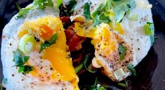 Two fried eggs with runny yolks are garnished with fresh herbs and chopped spring onions, served on a dark plate. A fork cuts into one egg, letting the yolk flow out over sautéed vegetables underneath.