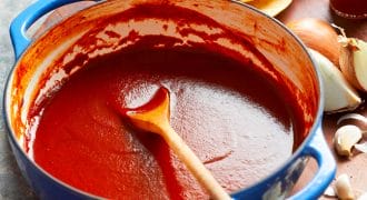 A blue pot filled with smooth red enchilada sauce sits on a wooden surface, surrounded by a wooden spoon, dried chillies, corn tortillas, a halved onion, garlic cloves, and a jar of hot sauce.