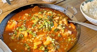 A frying pan filled with a red tomato-based stew topped with chopped spring onions sits on a wooden table, alongside a bowl of white rice, a salad, and empty plates with cutlery.