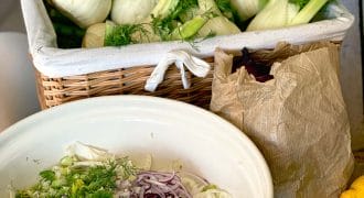 A wicker basket filled with fresh fennel bulbs sits behind a bowl containing sliced fennel, red onion, and herbs. A brown paper bag and two lemons are also on the table.