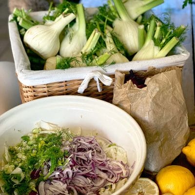 A wicker basket filled with fresh fennel bulbs sits behind a bowl containing sliced fennel, red onion, and herbs. A brown paper bag and two lemons are also on the table.