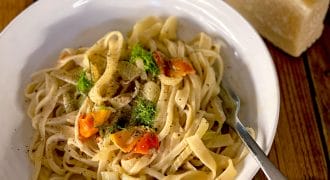 A white bowl of fettuccine pasta with cherry tomatoes, fresh herbs, and black pepper. A fork rests on the bowl, and a block of cheese with a grater is in the background on a wooden table.
