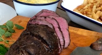 Sliced roast beef on a wooden chopping board with a knife, next to a bowl of chips and a bowl of yellow sauce with herbs, all set on an outdoor wooden table.