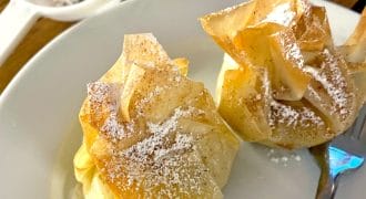 Two golden-brown pastry parcels dusted with icing sugar sit on a white plate with a fork; a mesh sieve with sugar is in the background on a wooden table.