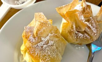 Two golden-brown pastry parcels dusted with icing sugar sit on a white plate with a fork; a mesh sieve with sugar is in the background on a wooden table.