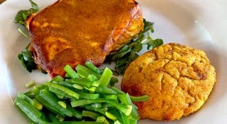 A white plate with a serving of lasagne on leafy greens, a scoop of sautéed green beans, and a golden-brown scone. The food is arranged neatly on a wooden table.