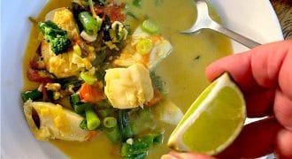 A hand squeezes a lime wedge over a bowl of fish and vegetable stew in a yellow broth, with a spoon resting inside the white bowl.