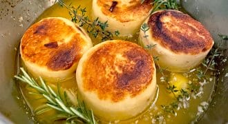 Four golden-brown, round potato slices are seared in a pan with melted butter, garnished with sprigs of fresh rosemary and thyme.