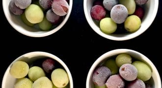 Four white bowls filled with green and purple grapes, some with a frosty appearance, arranged in a square pattern on a black background.