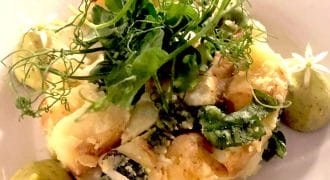 A plated dish featuring a creamy vegetable medley garnished with fresh herbs and edible flowers, accompanied by three dollops of green sauce, each topped with a small white flower.