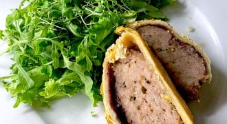 A plate with a leafy green salad and a sliced portion of meat wrapped in golden puff pastry, resembling a homemade pork Wellington, on a white plate.