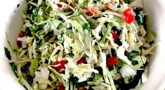 A white bowl filled with shredded cabbage salad mixed with chopped tomatoes, coriander, and green herbs, placed on a wooden surface.