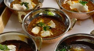 Six metal bowls filled with spicy red curry, garnished with herbs and chunks of white fish, are arranged on a tray. The curry has a rich, oily surface and each bowl has ornate metal handles.