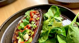 A dark plate with a roasted aubergine half topped with chopped spring onions, red chilli, and herbs, served alongside a fresh green salad.