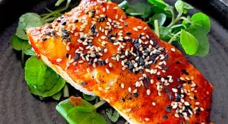 A fillet of salmon topped with a red glaze and garnished with white and black sesame seeds, served on a bed of fresh green leaves on a dark plate.