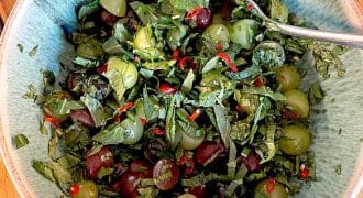A bowl containing chopped green herbs, red and green grapes, sliced red chilli, and black olives, with a spoon resting in the mixture on a wooden surface.