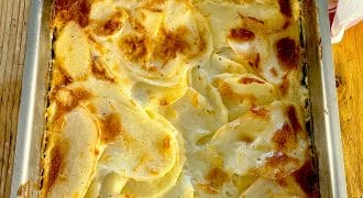 A baked potato gratin in a rectangular metal dish, featuring thinly sliced potatoes layered with cream and cheese, with a golden-brown, slightly crispy top. The dish sits on a wooden surface.