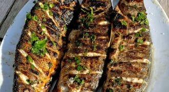 Three grilled whole fish with char marks and garnished with chopped herbs are served on a white oval plate, set on a rustic wooden table in bright sunlight.