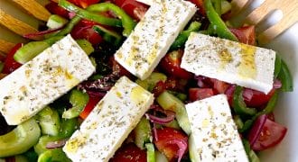 A bowl of Greek salad with sliced tomatoes, cucumbers, green peppers, red onions, and five rectangular pieces of feta cheese, garnished with dried herbs and olive oil. Wooden salad servers rest at the side.